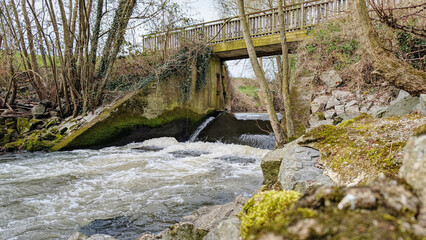 Flowing stream beneath wooden pedestrian and bike bridge surrounded by rocky terrain and greenery in a tranquil rural setting in France.