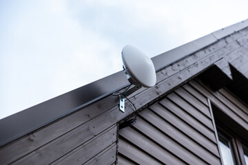 A satellite dish style WiFi router mounted near the roofline of a dark wooden house with sloped roof, set against an overcast sky.