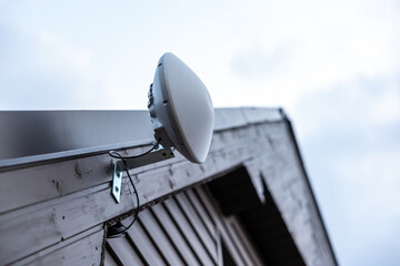 A white dome shaped Wi Fi router is mounted on the roof edge of a wooden house. Cables are neatly...
