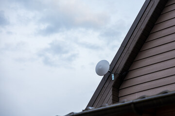 Upper section of a private house with wooden siding, steep roof, and a white satellite dish mounted on the side. Partially cloudy sky in the background.