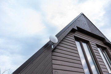 Upper section of a private house with wooden siding, large vertical windows, and a satellite dish on the slanted roof under an overcast sky.