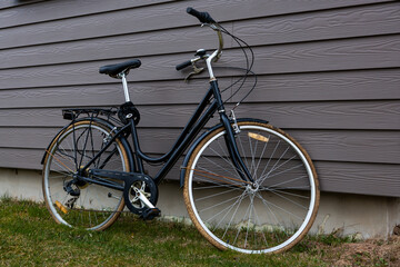 A black bicycle with a step through frame leans against a gray wooden house wall. Grass and soil are visible at the base, suggesting a residential setting.