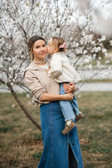 Fototapeta premium Stylish woman wearing denim skirt and beige sweater holding baby girl 2-3 year old over flowers in park. Motherhood.