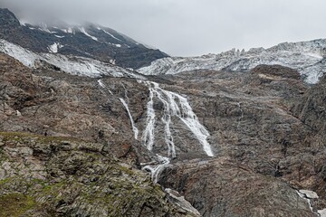 Rushing waters tumble gracefully over steep, jagged rocks in North Ossetia, creating a stunning display of nature's power. The atmosphere is dramatic, with cloudy skies enhancing the scene