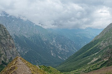 Naklejka premium Lush greenery hugs the rugged mountains of North Ossetia, where dramatic clouds swirl above. The landscape showcases the beauty of nature's contrasts in this serene and captivating area