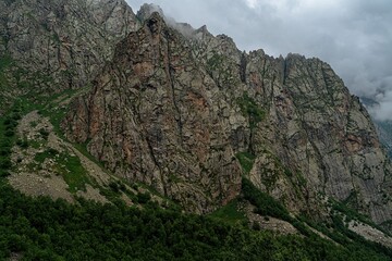Towering granite cliffs in North Ossetia create a breathtaking backdrop, shrouded in mist. The lush greenery below contrasts with the rugged rock formations, evoking a sense of serenity
