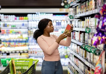 Fototapete Pretty African American woman choosing groceries at supermarket, shopping for food, buying products for her family. Female consumer purchasing goods at huge mall, walking among shelves © Prostock-studio