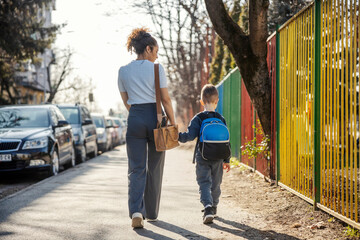Rear view of young mother holding hands with her son and taking him to kindergarten.