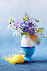 Easter still life with a bouquet of spring flowers in an eggshell on an egg stand, feathers and decor on a white and blue background, beautiful card