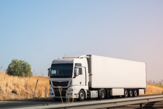A large European trailer truck drives along a road with a grain field next to it. The truck is white to be able to make a mockup with perosonalized text or logo