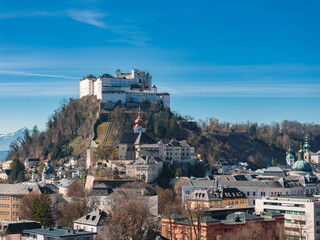 Fototapeta premium Aerial view of Salzburg, Austria, featuring Hohensalzburg Fortress on a hill, a red spired church, traditional buildings, and snow capped mountains.