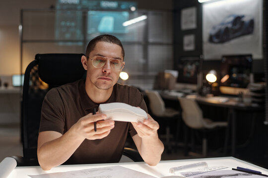 Young serious development engineer sitting by workplace in car design bureau and examining new hybrid crossover prototype