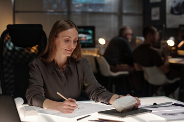 Young smiling woman looking at model of car and making sketch on paper while working over new automobile item