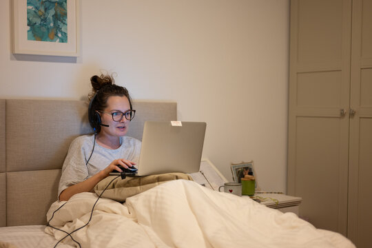 Young woman wearing headset working from home using laptop in bed, early in the morning or late at night