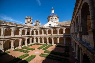 Obraz premium Historic courtyard with symmetrical arched walkways and lush greenery set beneath a clear blue sky in Valencia, featuring a striking dome and architectural details.