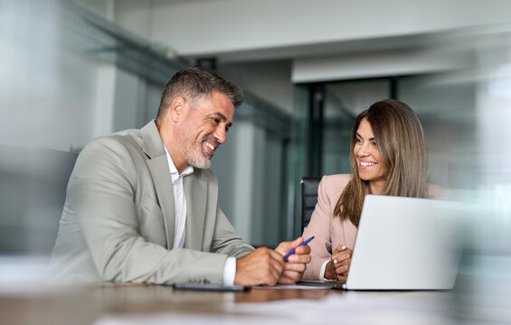 Happy professional mature businessman and businesswoman executive colleagues of middle age in suits sitting at table having partnership business discussion using laptop at corporate meeting in office.