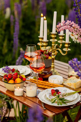 Simple countryside beautiful wedding decor, surprise romantic date outdoors, garden arrangement with lupins flowers on a meadow. Fruits, rose wine, burning candles. Sunset, candlelight, golden hour.