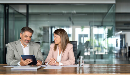 Fototapeta premium Two busy middle aged colleagues company board discussing smart technology at corporate meeting. Professional business executives office team working using digital tablet computer sitting at table.