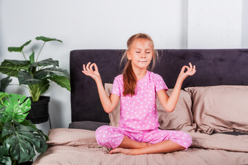 Little beautiful girl meditating in lotus position sitting on bed in cozy bedroom. Healthy lifestyle. Hobbies.