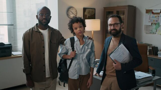 Dolly out group portrait of bearded Caucasian psychologist with clipboard posing with African American troublemaker schoolboy with bandaged nose and torn shirt and his dad indoors, looking at camera