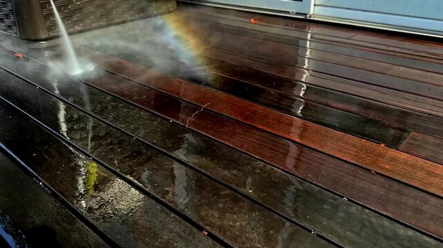 A person is using a pressure washer to clean a wooden deck in a suburban backyard. The sunlight reflects off the water as dirt and grime are removed, revealing the clean surface beneath