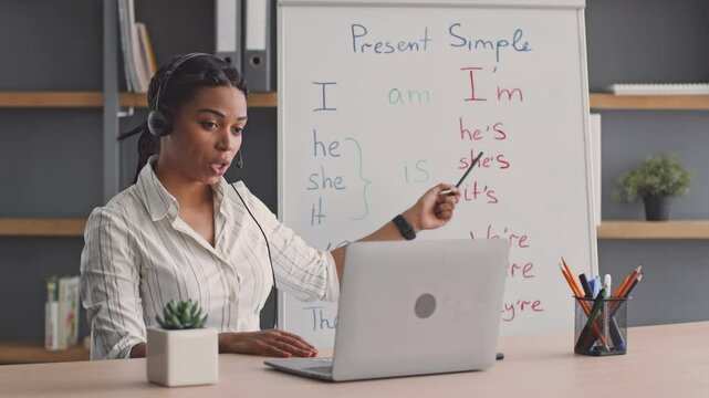 A young African American woman teaches English grammar online, engaging her students with clear explanations. She uses a whiteboard and smartphone camera to enhance her lesson.