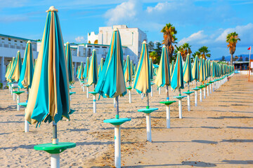 Sandy beach with a neat row of closed, colorful umbrellas. Background with building and palm trees...