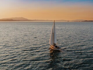 A sailboat with three people aboard glides across the water at sunset in  San Francisc, California,...
