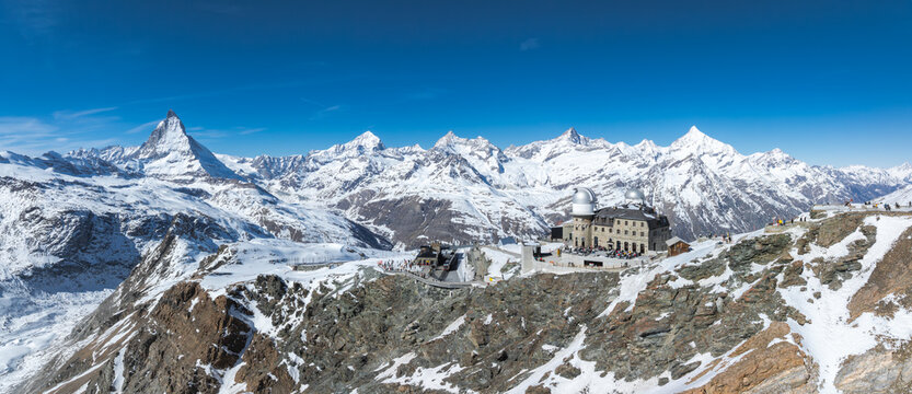 Snow covered Swiss Alps with Matterhorn on the left, Gornergrat Observatory in the foreground, ski trails, and visitors under a clear blue sky.