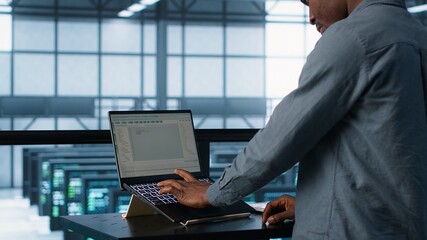 Computer scientist in server room coding on laptop, developing disaster recovery plans. African...