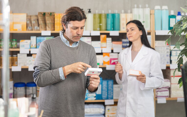 Adult man buyer choosing box of paracetamol tablets with help of adult woman pharmacist in pharmacy