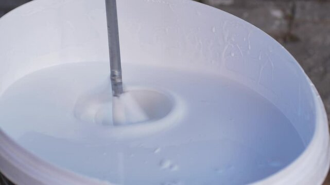 Plaster mixer or universal tool lowers into container with white paint, stirring for even texture. This step prepares paint for smooth application on walls during home or building construction