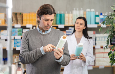 Adult man buyer choosing shampoo with help of adult woman pharmacist in pharmacy