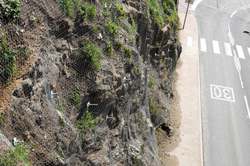 Workers implement netting and other methods to stabilize a rocky hillside along a roadside.