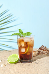 Glass of cold Cuba Libre cocktail and seashells on sand near blue background