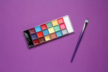 Face paints and brush on purple background, top view