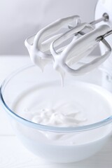 Hand mixer with whisks and bowl of whipped cream on white wooden table, closeup