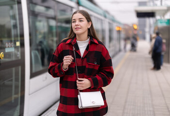 Young woman stands at a public transport stop and waits for a tram to go to a meeting. Girl uses public transport for a trip © JackF