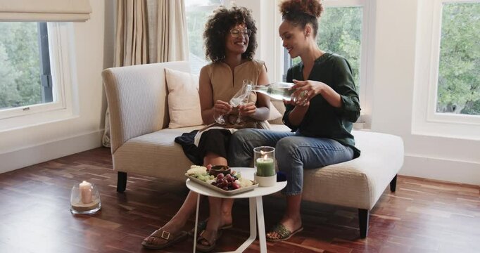 Toasting with champagne, same-sex diverse female couple relaxing on sofa together, at home