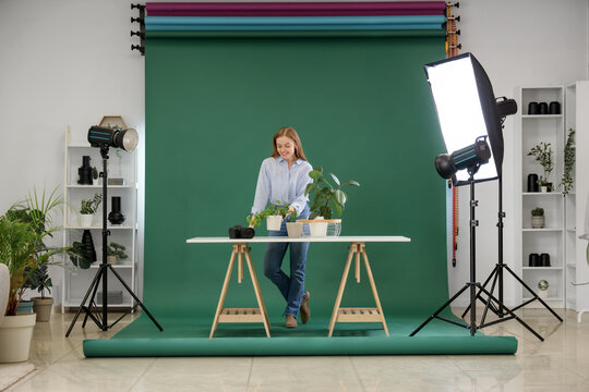 Female photographer with plants on table near green backdrop in photo studio