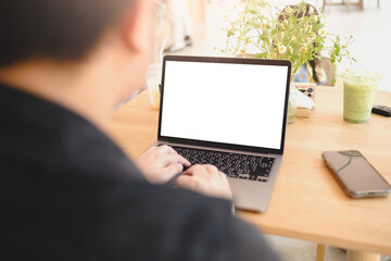 Back view of a man typing on a laptop with a blank white screen at a wooden café table, alongside a green iced drink and a smartphone.