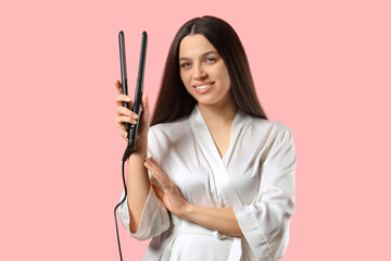Pretty young woman in bathrobe with hair straightener on pink background
