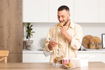 Young man with mobile phone eating paw shaped cookie in kitchen