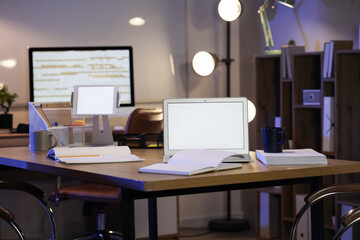 Blank laptop with tablet computer and notebooks on desk in office at night