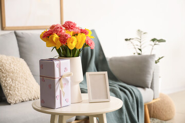Vase with flowers, blank frame and gift for Mother's Day on table in living room