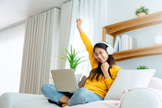 Young happy lucky woman student feeling excited winning, using computer laptop and sitting on sofa, adorable Asian female receiving great news on notebook, getting new job celebrating success