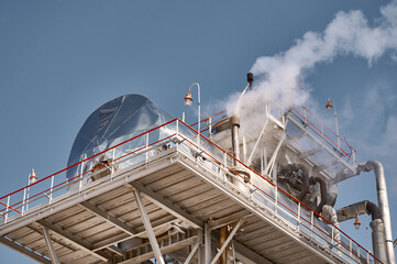 A distillation column at a chemical plant releasing steam under a clear sky, showcasing industrial operations in a vibrant setting