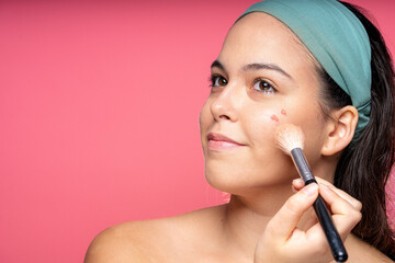 Young woman applying blush with makeup brush on pink background