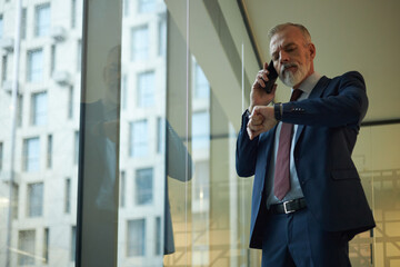 Low angle view of mature Caucasian man checking time on wrist watch while talking on phone, standing in modern office with panoramic windows