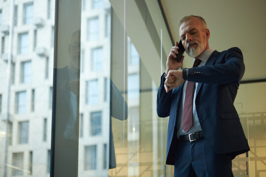 Low angle view of mature Caucasian man checking time on wrist watch while talking on phone, standing in modern office with panoramic windows
